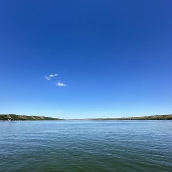 This photo was taken in the middle of Katepwa Lake, Saskatchewan. It depicts the lake framed by the hills of the valley, all under a blue, almost cloudless sky.